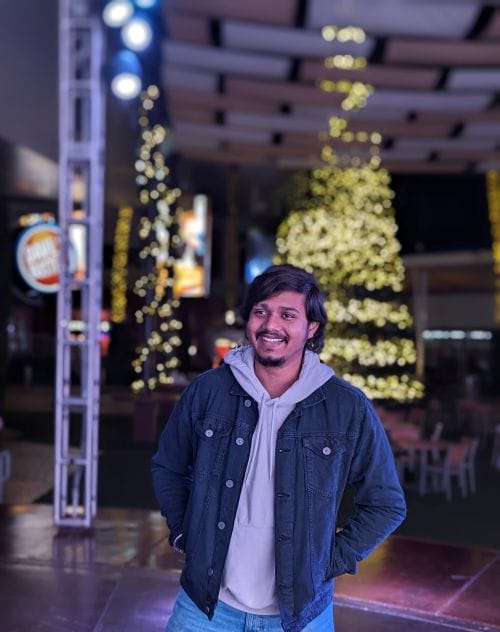 A smiling young man wearing a light hoodie, dark denim jacket, and blue jeans, standing in front of illuminated Christmas trees at night.
