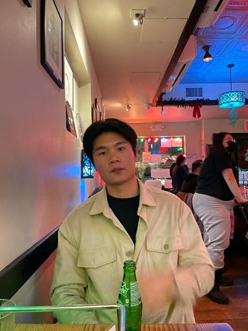 A young man sitting at a table inside a dimly lit restaurant with a green soda bottle in front of him.