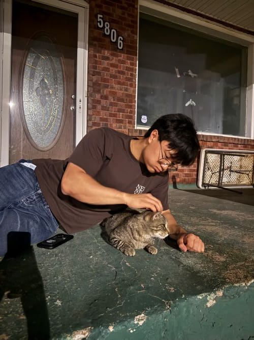 A young man lying on a concrete porch at night, gently petting a tabby cat resting beside him.