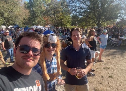 Three young men posing together outdoors at a crowded sunny outdoor event, with one wearing sunglasses taking the selfie.