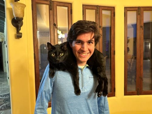 A smiling young man wearing a light blue quarter-zip pullover with a sleek black cat draped across his shoulders.